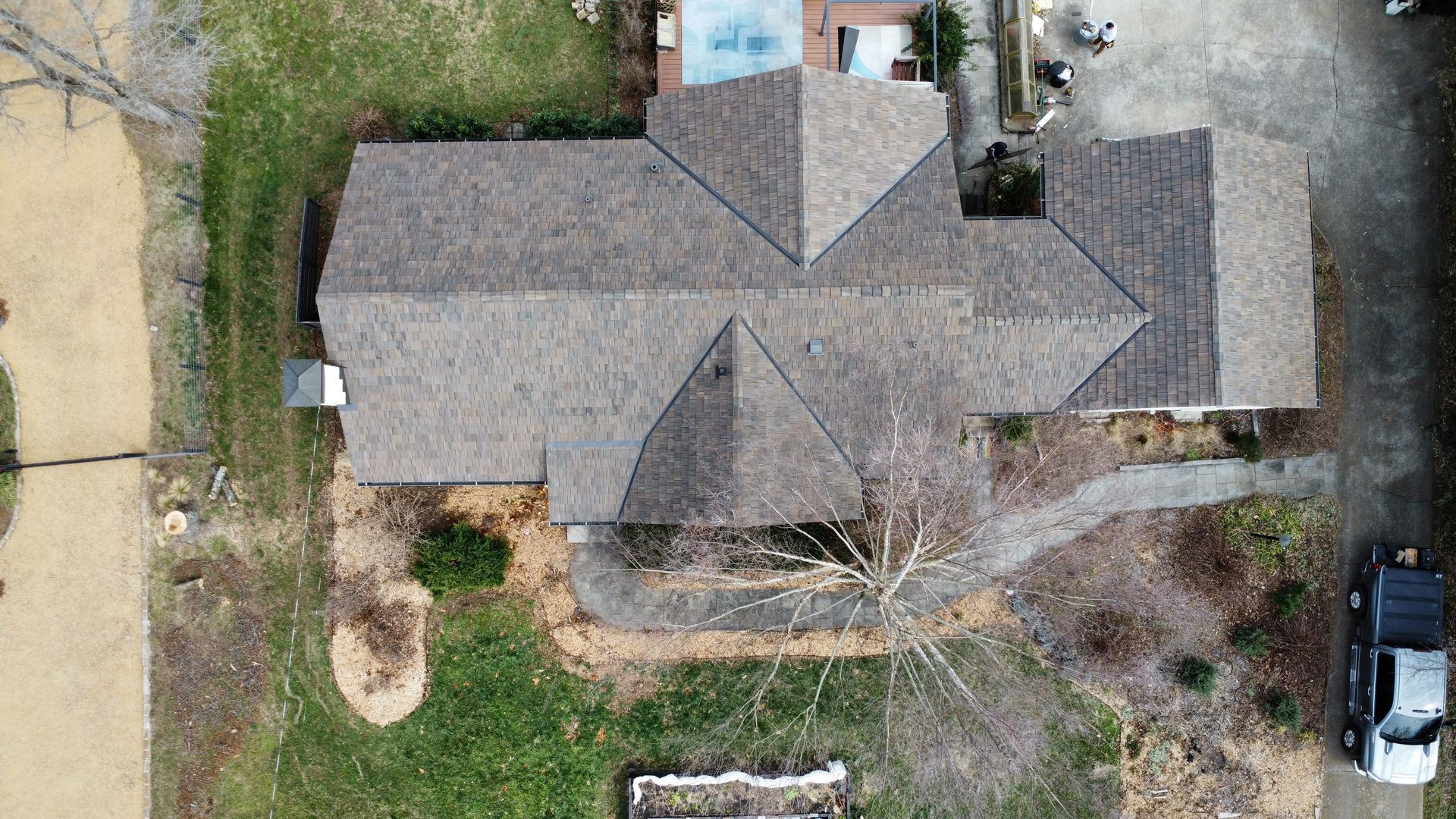 Aerial view of a house with a gray roof, yard, driveway, and parked vehicles beside it.