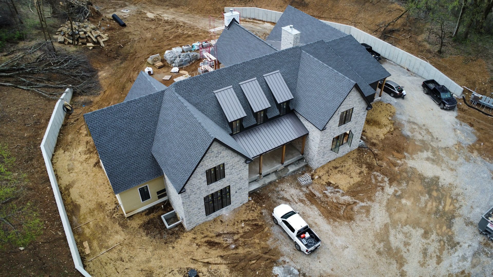 Aerial view of a large gray-roofed house with solar panels, surrounded by dirt and parked vehicles.