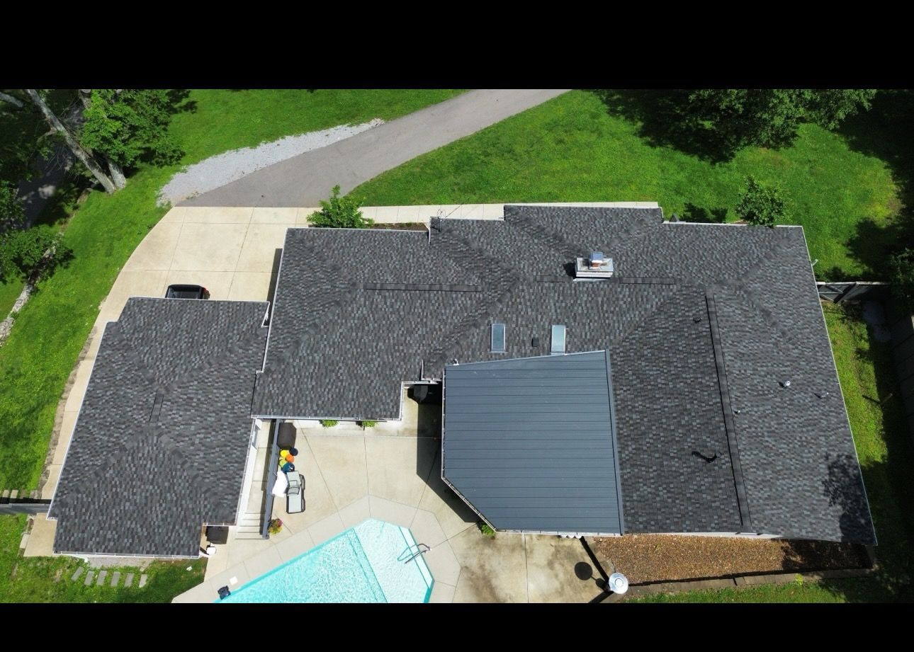 Aerial view of a house with a dark shingled roof, backyard patio, driveway, and green lawn.