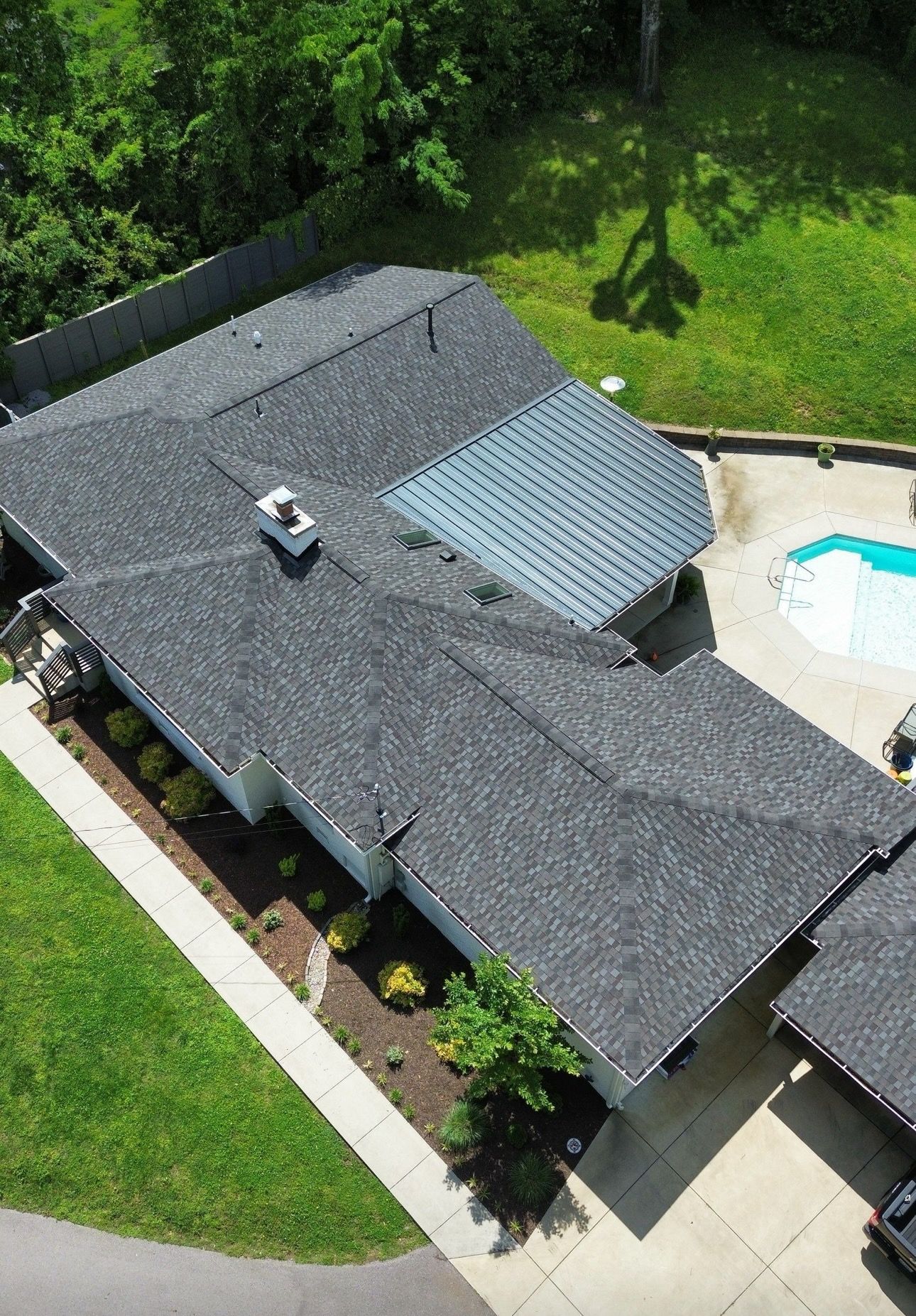 Aerial view of a dark shingled house with a pool and green lawn beside it.