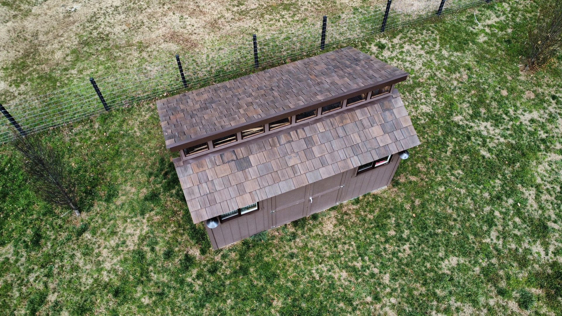 Aerial view of a small brown shed with a pitched roof in a grassy fenced area.