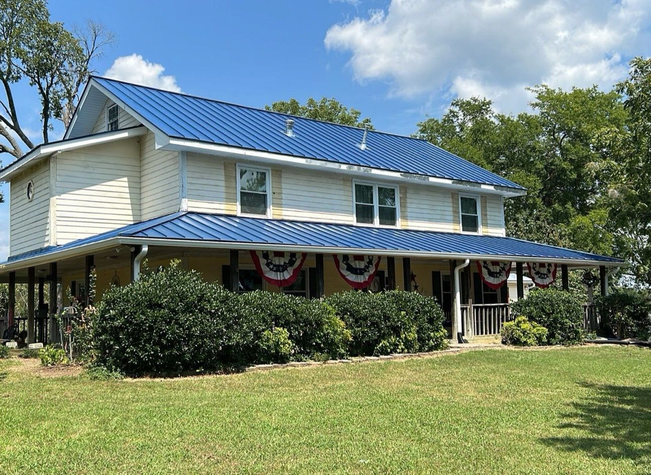 Two-story white house with blue roof and porch bunting on a grassy lawn under a clear sky