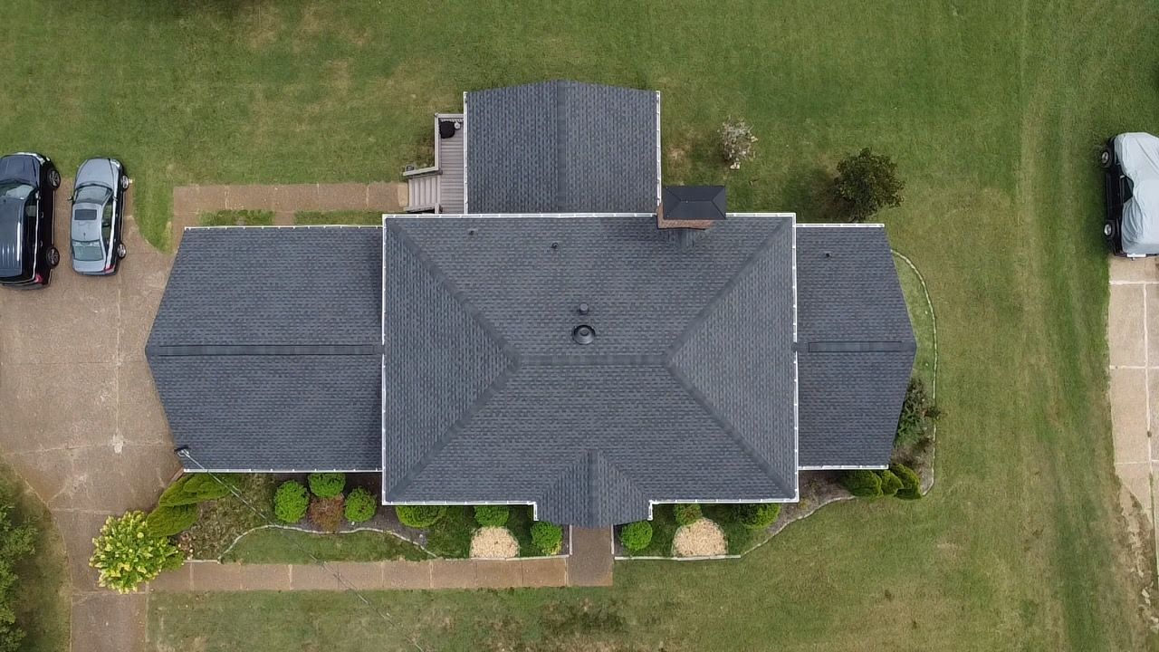 Aerial view of a gray-roofed house with a driveway, lawn, and parked cars.