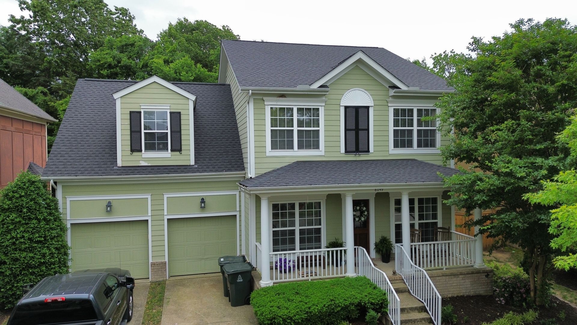 Two-story green house with white trim, front porch, garage, and black car in the driveway