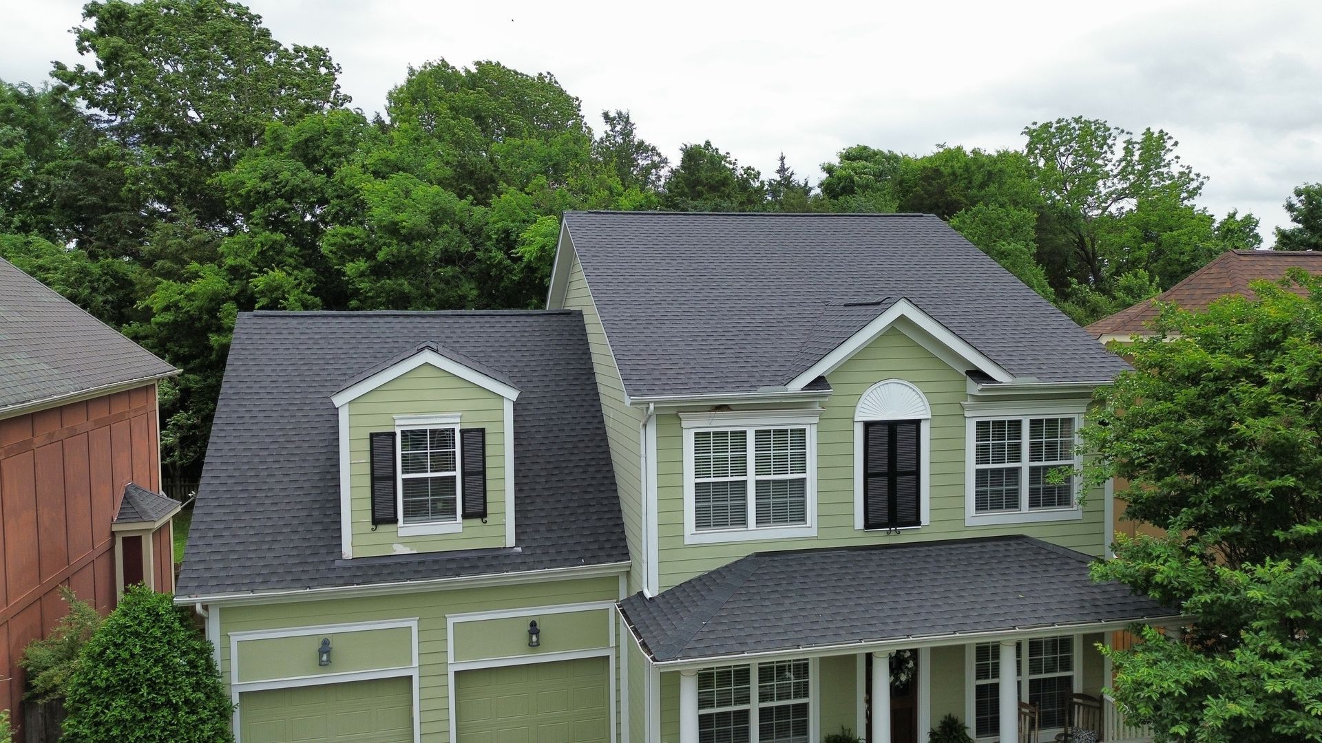 Aerial view of a green house with dark gray roofs surrounded by trees and neighboring homes