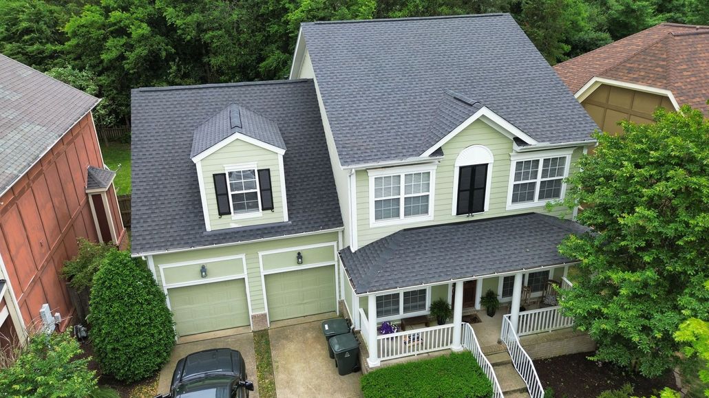 Aerial view of a gray two-story suburban house with white trim, garage, and front porch surrounded by trees