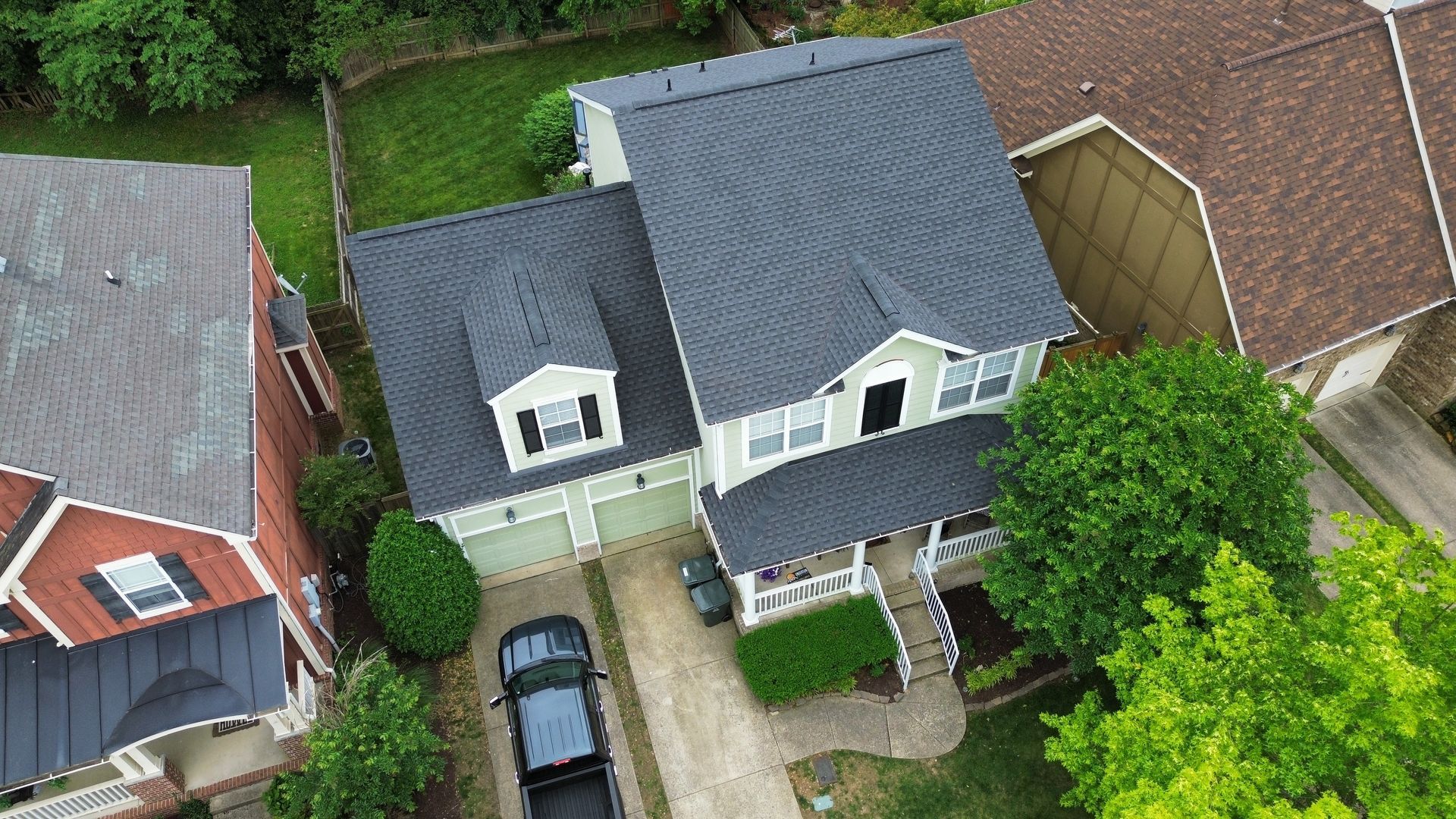 Aerial view of two suburban houses with gray roofs, driveway, parked car, and green trees