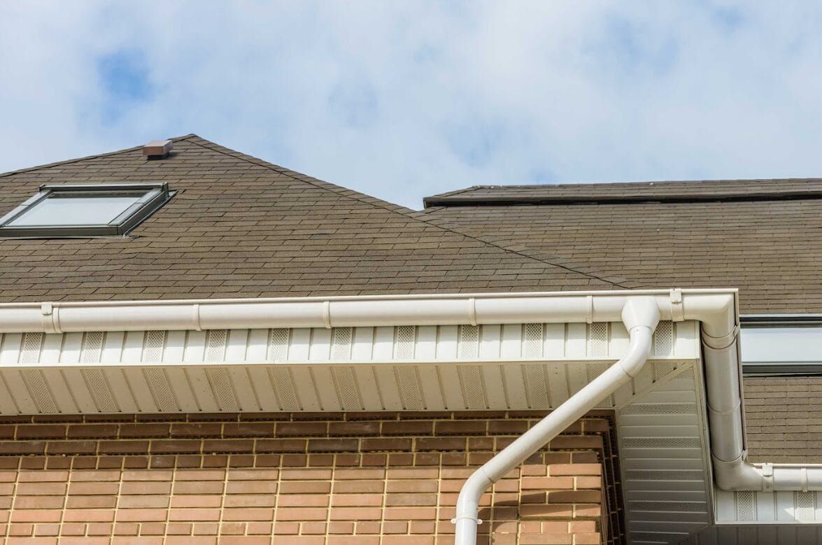 Brown shingled roofs with a skylight, white gutters, and brick exterior under a cloudy sky.