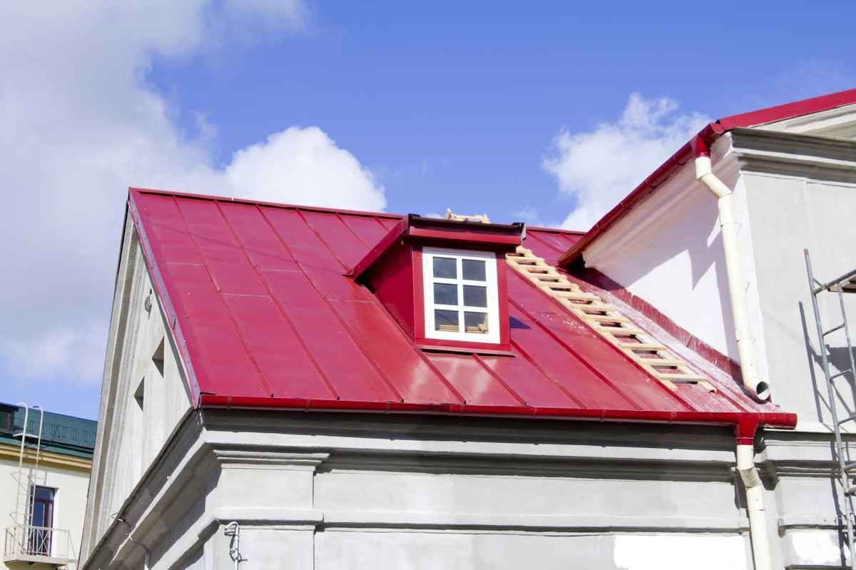 Red metal roof on a white building, with a dormer window against a blue sky.