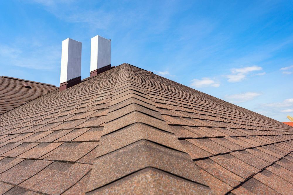 Brown shingle roof with two white chimneys against a bright blue sky.