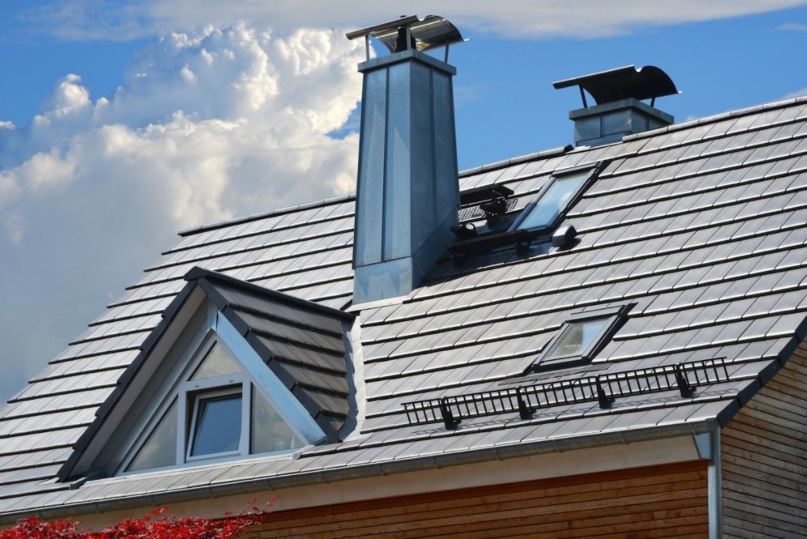Close-up of a house roof with a dormer window, chimney, and skylight against a cloudy blue sky.