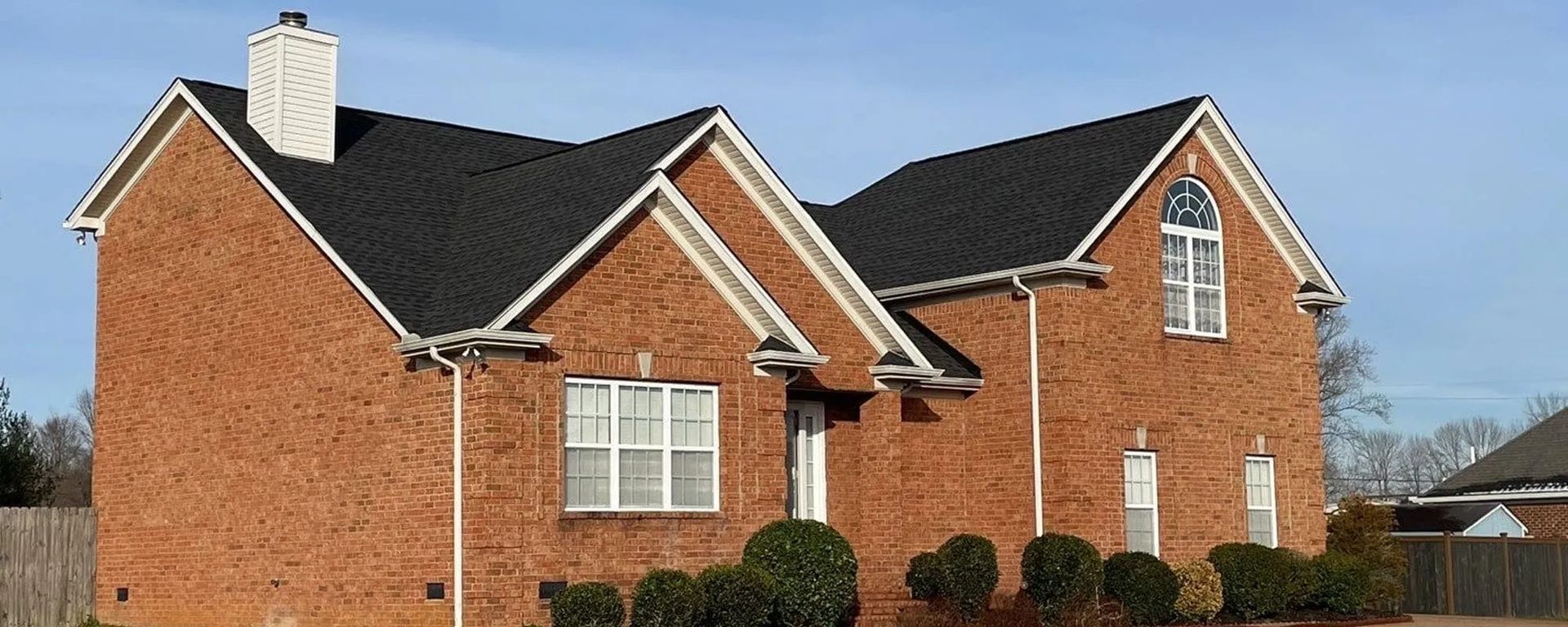Brick house with black roof, white trim, and small bushes in front. Blue sky in background.