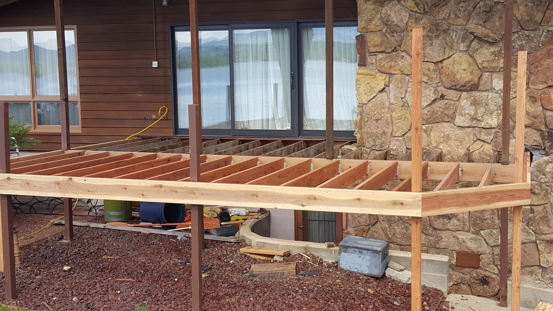 Partially built wooden deck beside a stone house with windows and a ladder underneath.