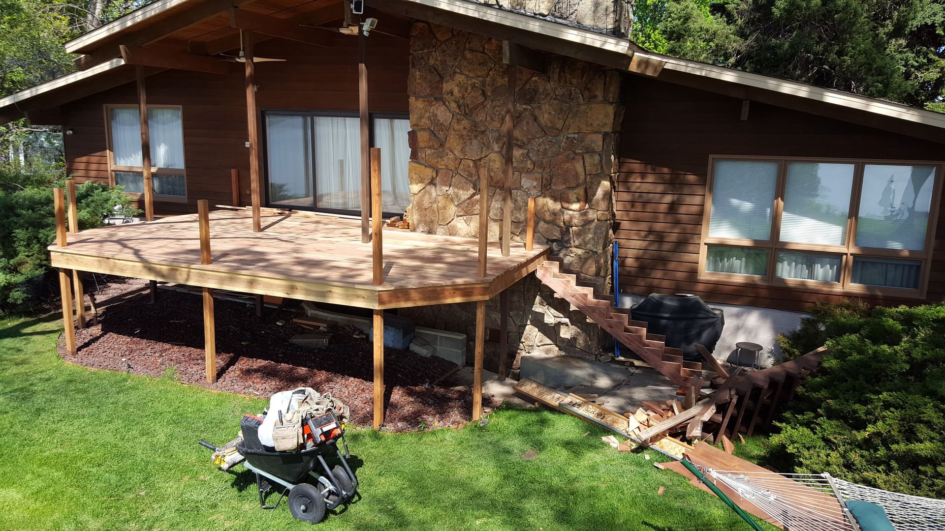 Rustic stone house with wooden deck and large windows beside a grassy yard, with a stroller in front.