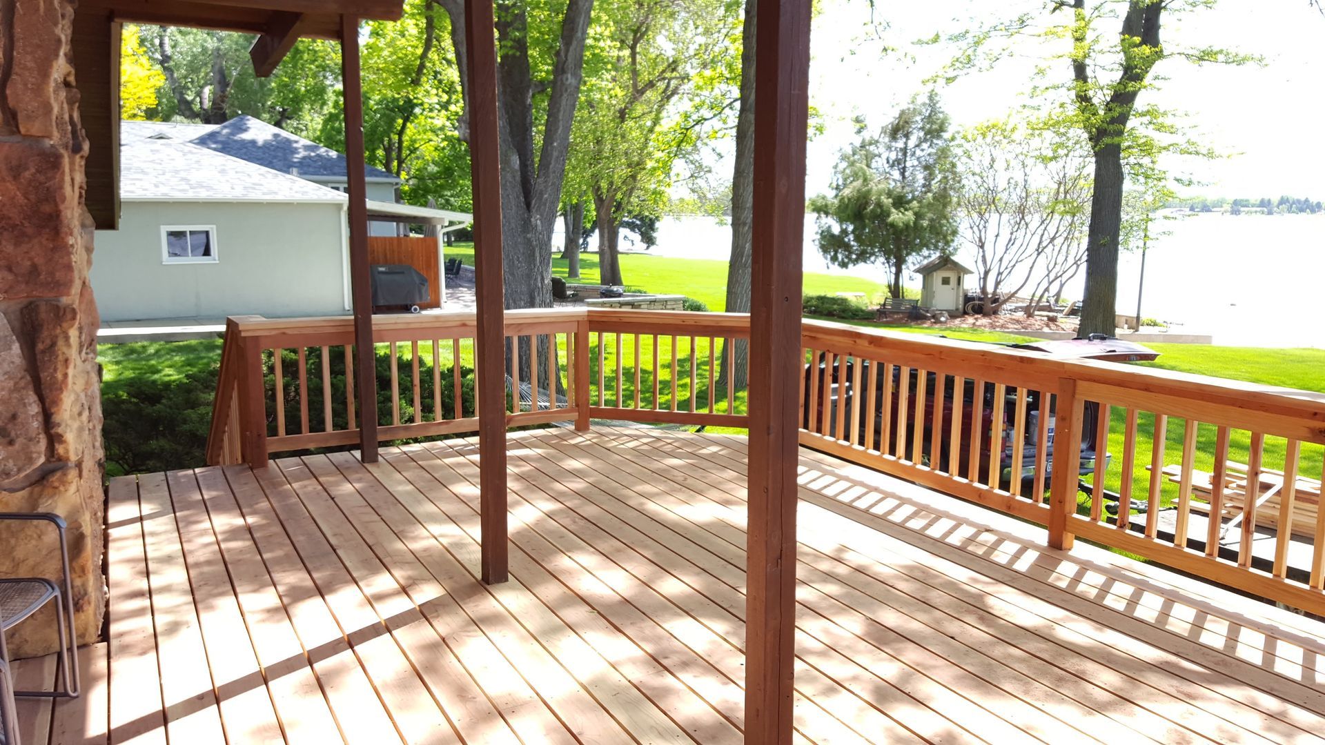 Wooden deck with railings under trees beside a lakeside yard and house.