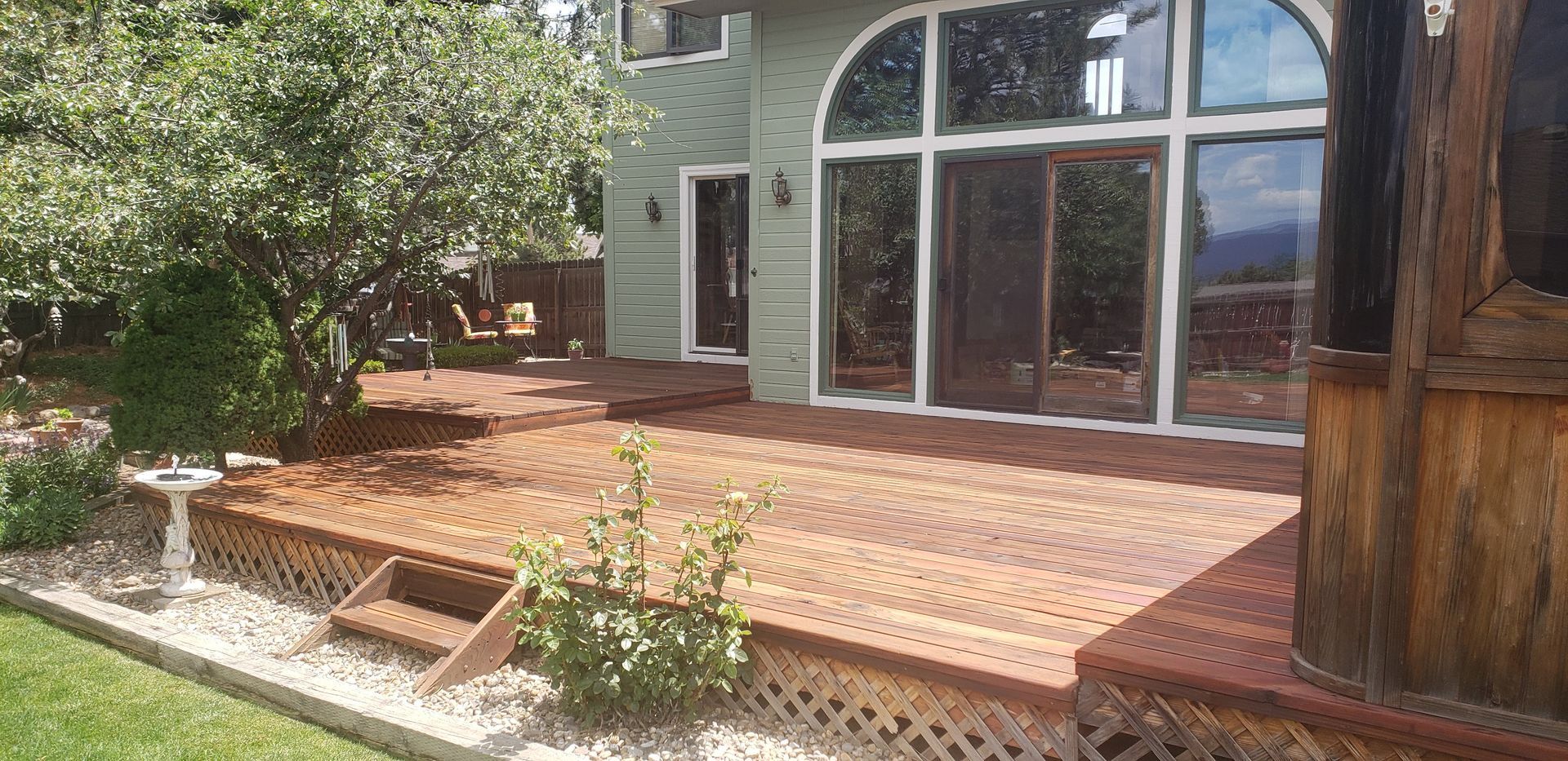 Wooden deck with steps leading to arched glass doors on a brick house, bordered by trees and landscaping.