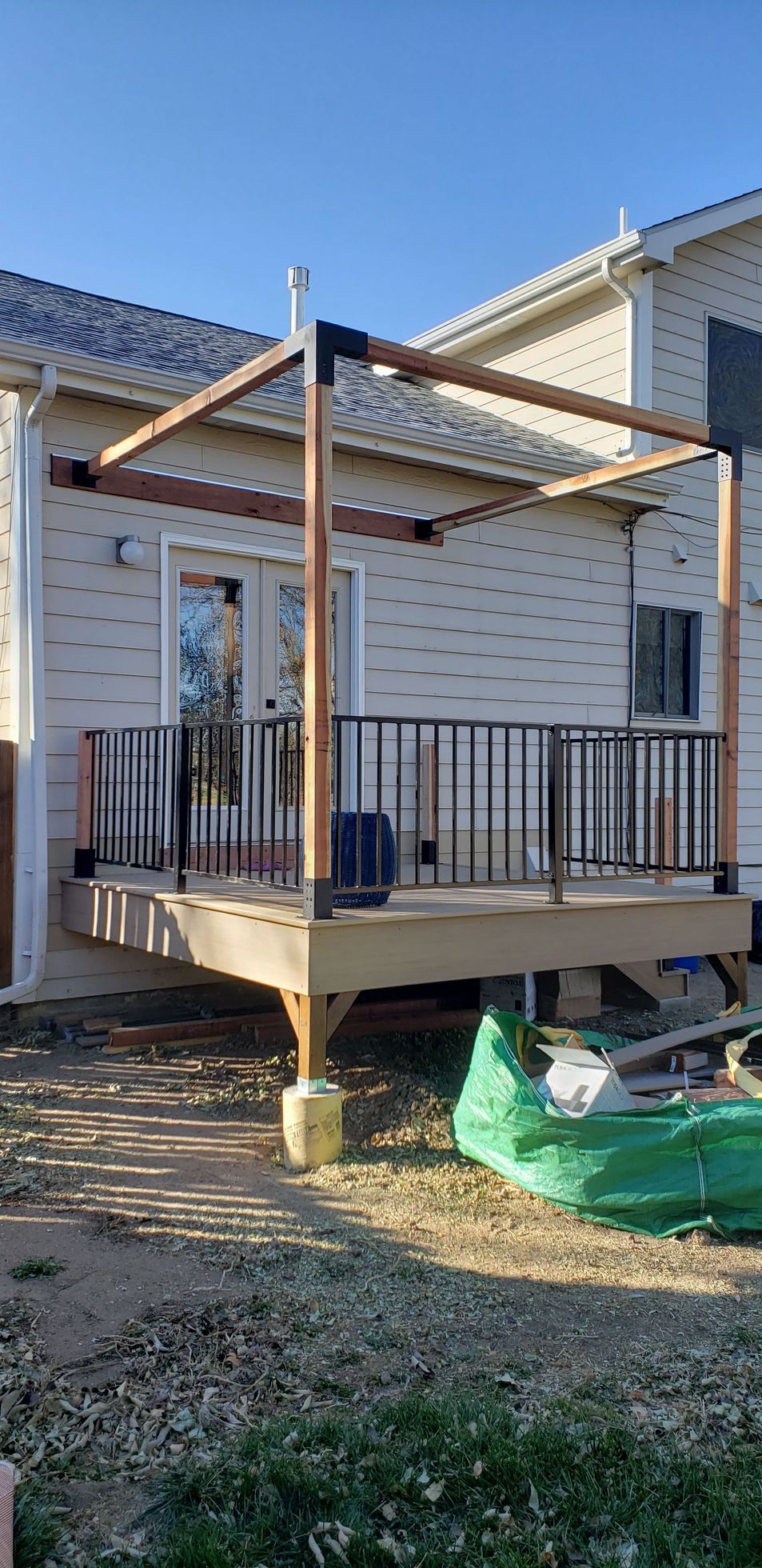 Back deck under construction beside a white house, with wooden posts, black railing, and a tarp on the ground.