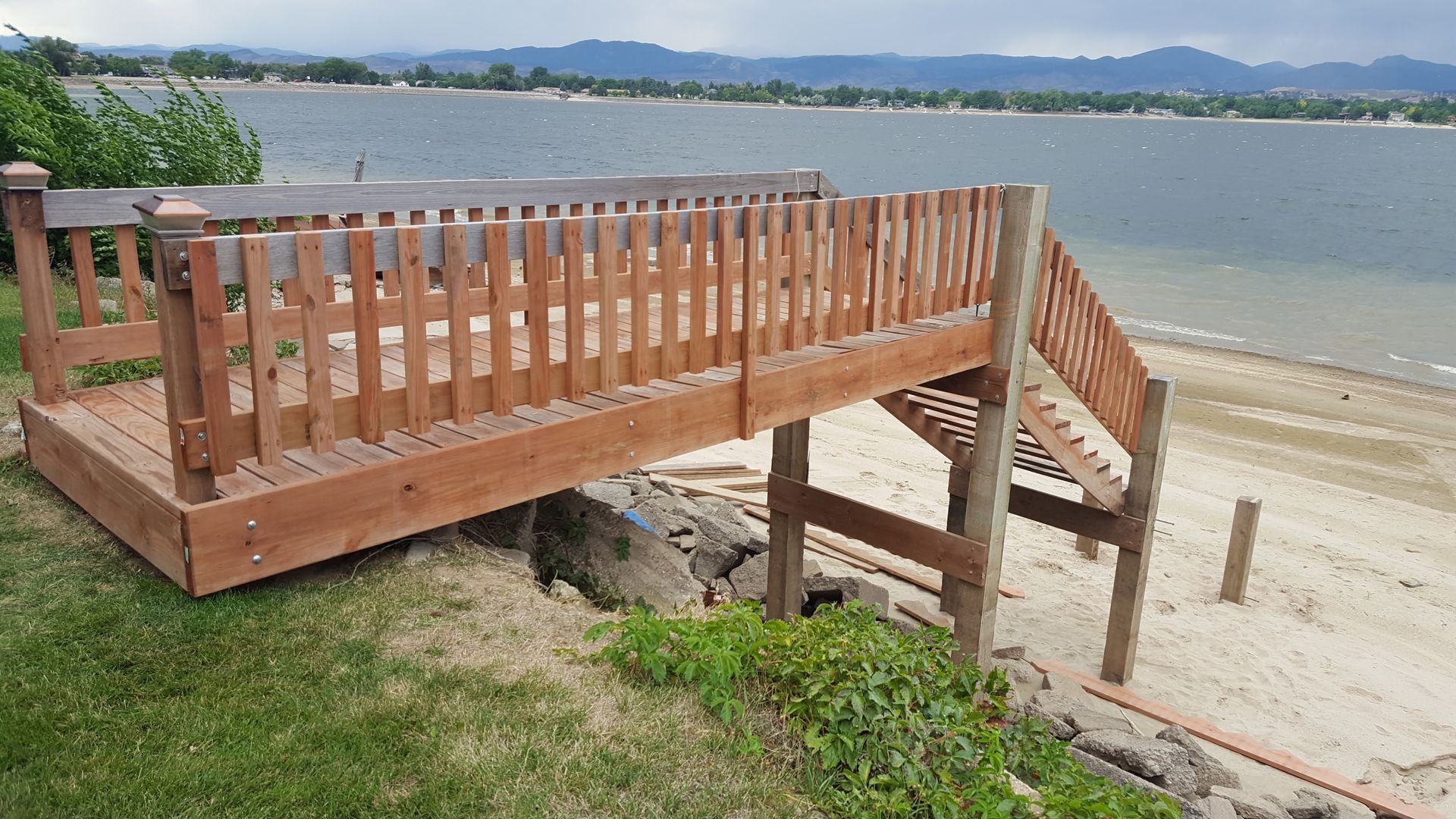 Wooden beach ramp with railing overlooking a sandy shore and calm water under a cloudy sky.