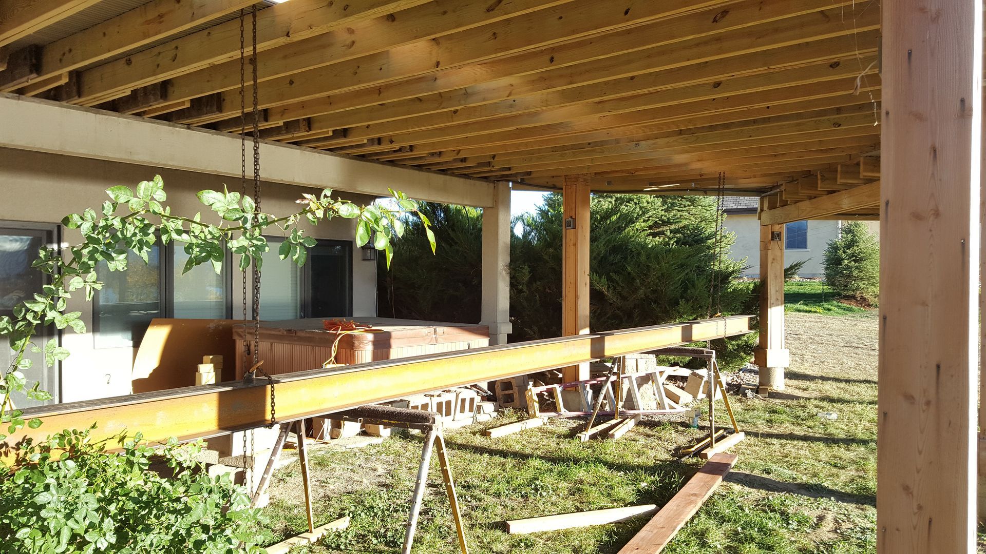 Covered wooden patio with posts, beams, and stacked lumber in a sunlit backyard construction area.