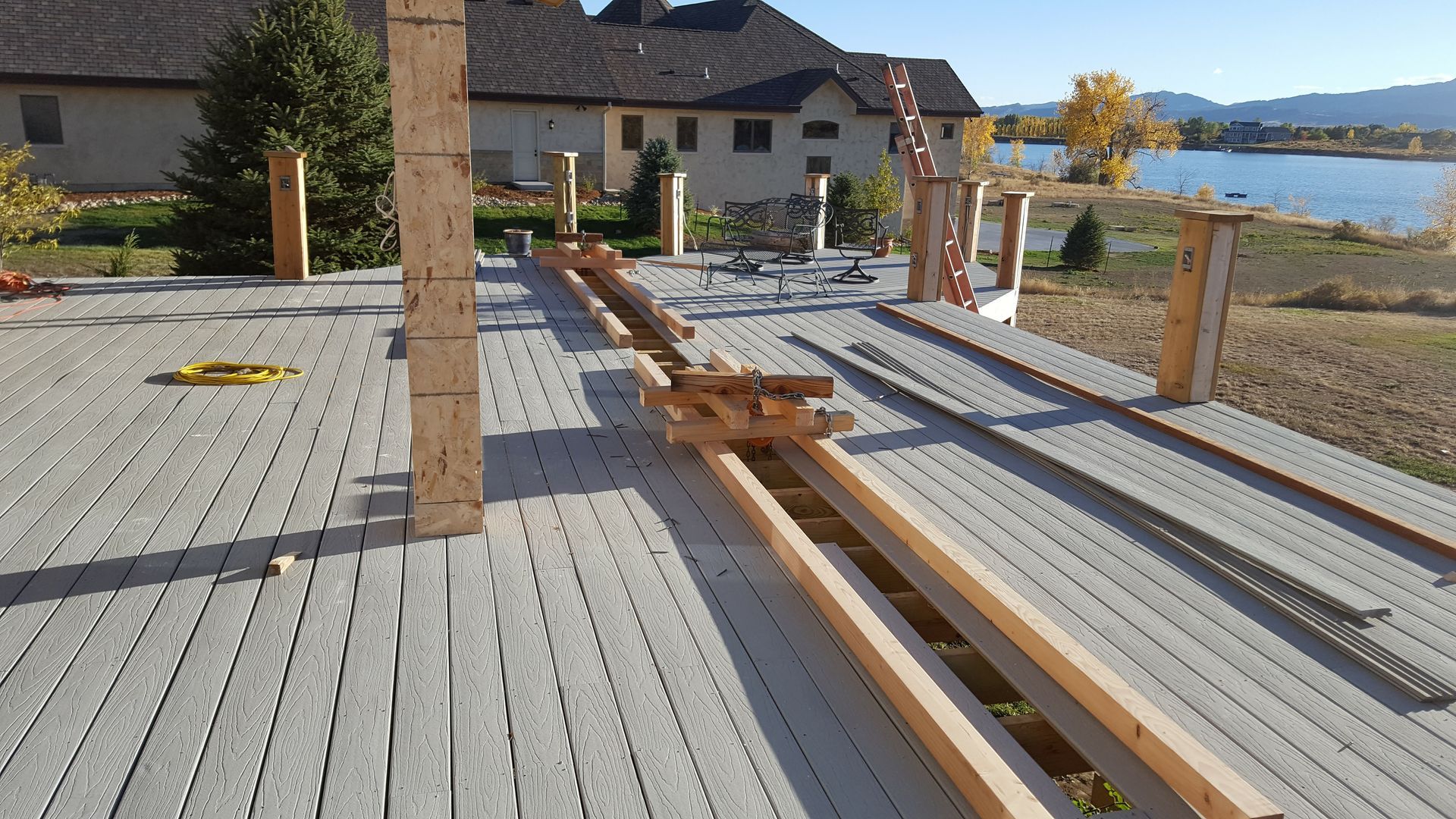 Wooden deck under construction with beams, posts, and a house by the water in the background.