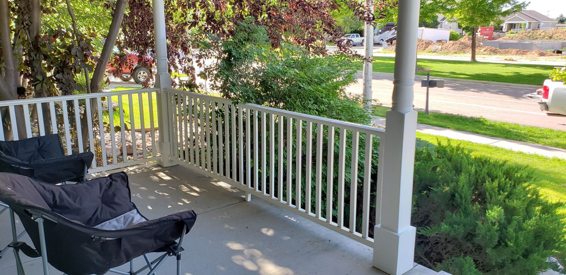 Front porch with two black folding chairs, white railing, and a sunny street view beyond the shrubs.
