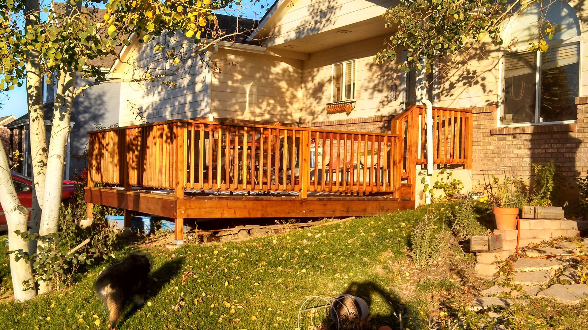Wooden front deck with railing on a sunlit house, surrounded by trees and autumn leaves.