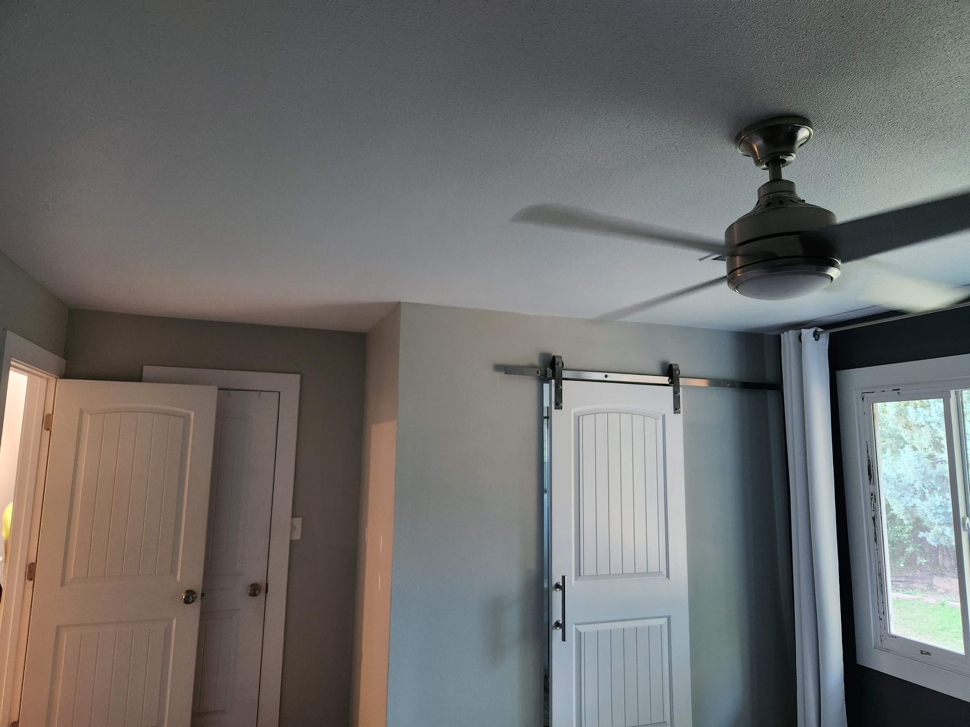 Bedroom with gray walls, white closet doors, ceiling fan, and a window with a dark curtain.