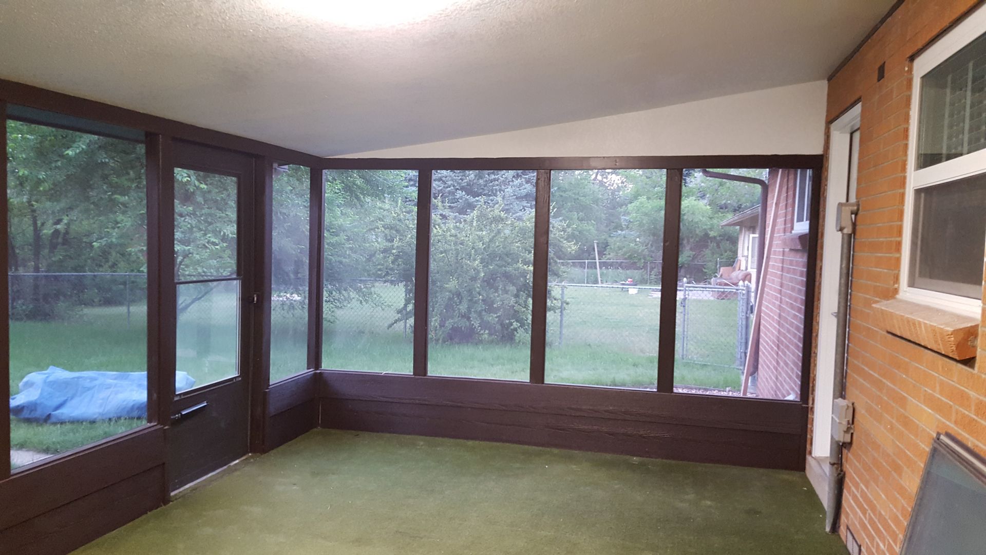 Enclosed screened porch with green carpet, wood-paneled wall, and large windows overlooking a yard.