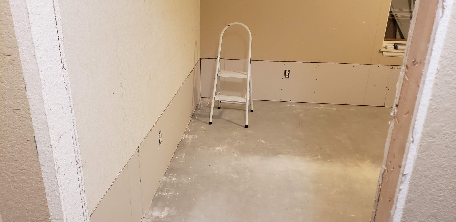 Empty unfinished room with beige walls, bare floor, and a small step ladder against the back wall.