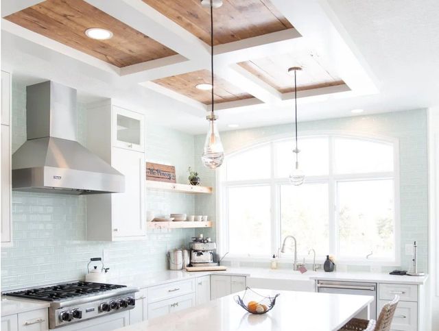 Bright modern white kitchen with wooden ceiling accents, stainless range hood, island, and sunlit windows