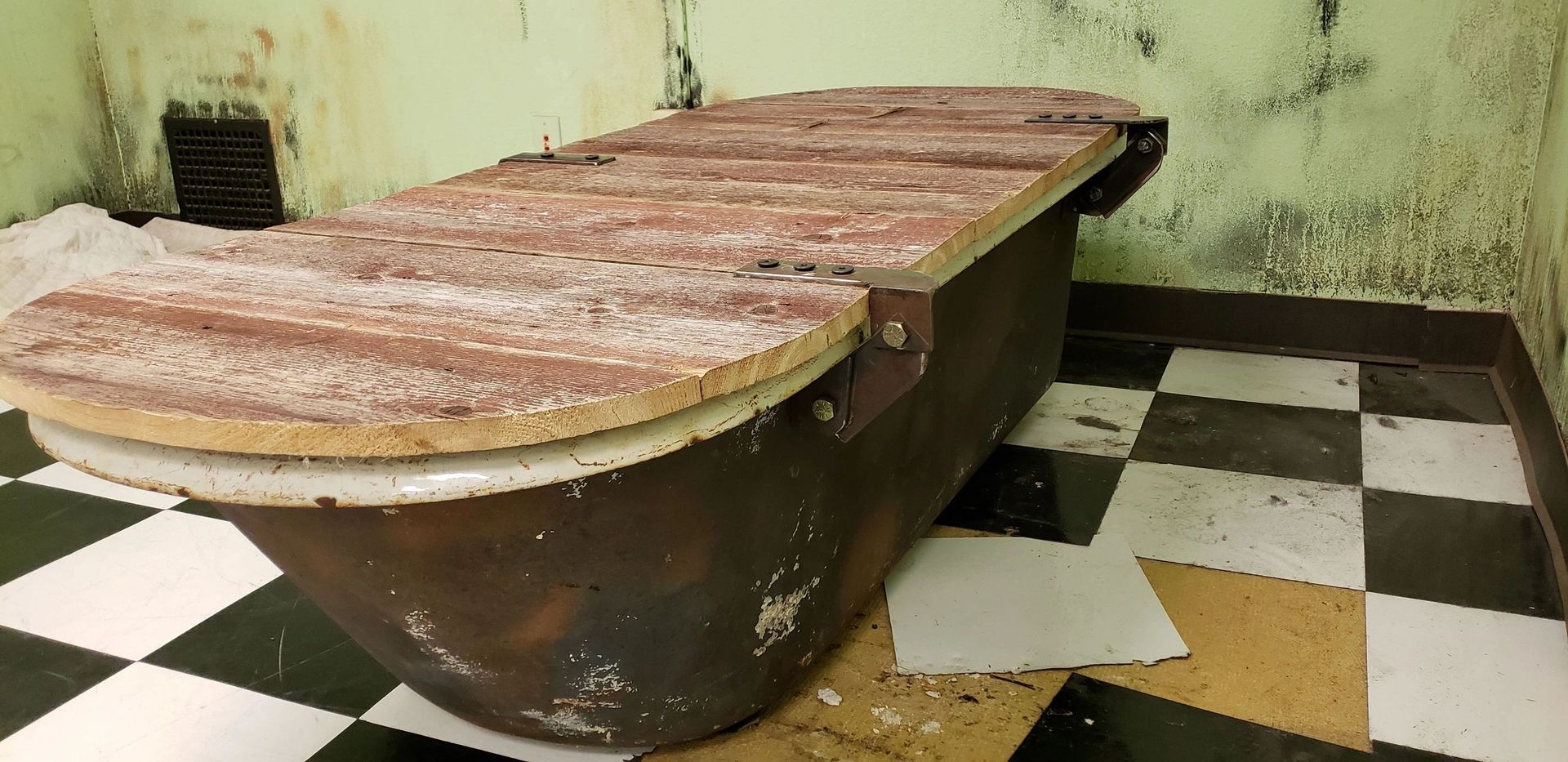 Old wooden bathtub or trough in a worn room with black-and-white checkered tile floor.