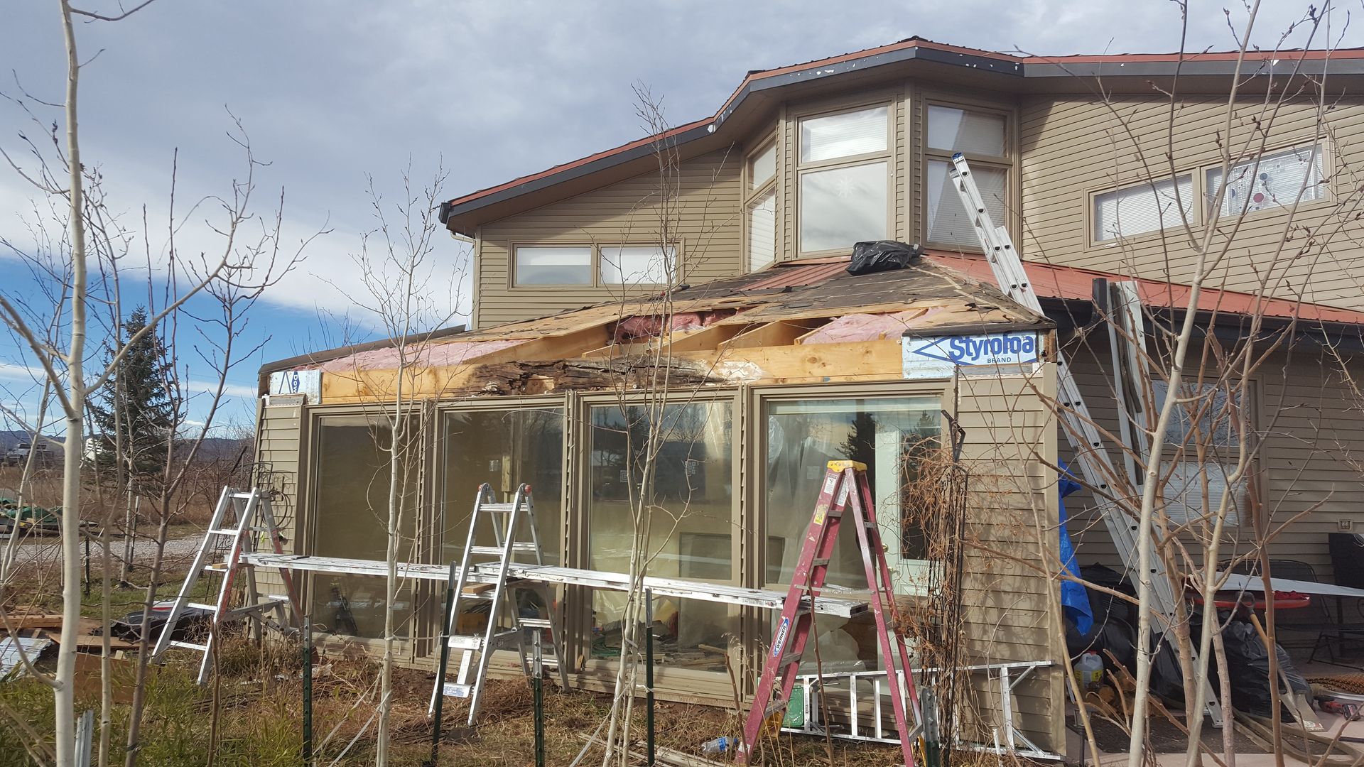 Partially demolished house with exposed roof framing and construction ladders in front.
