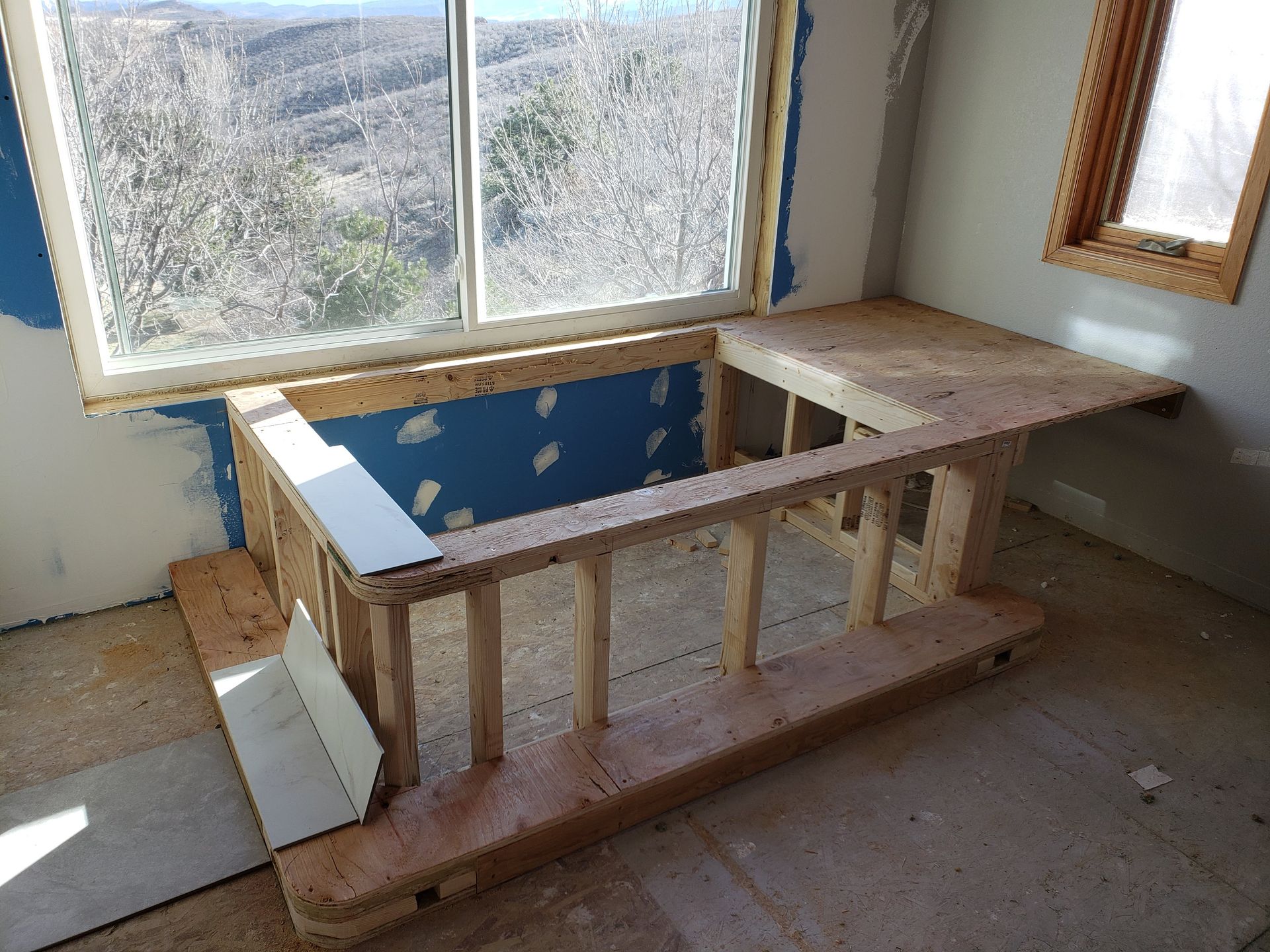 Unfinished built-in bathtub frame under construction beside large windows in a room with exposed studs.