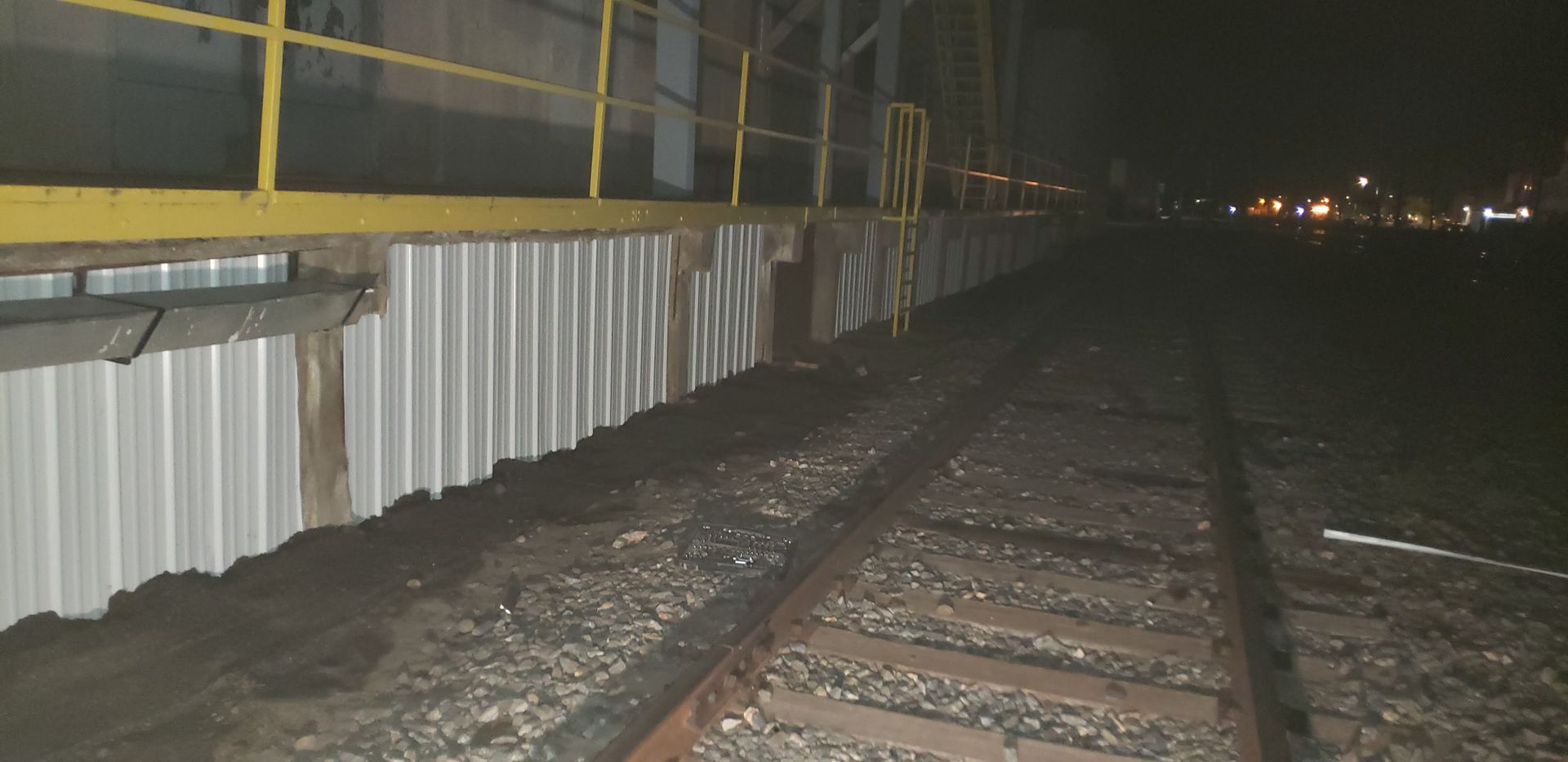 Nighttime view of railroad tracks beside a lit industrial fence and gravel embankment.