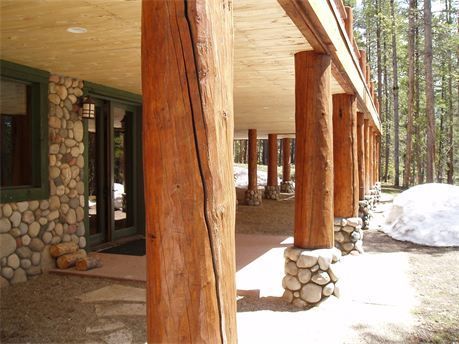 Rustic lodge porch with log columns, stone walls, and snow beside a forested walkway.