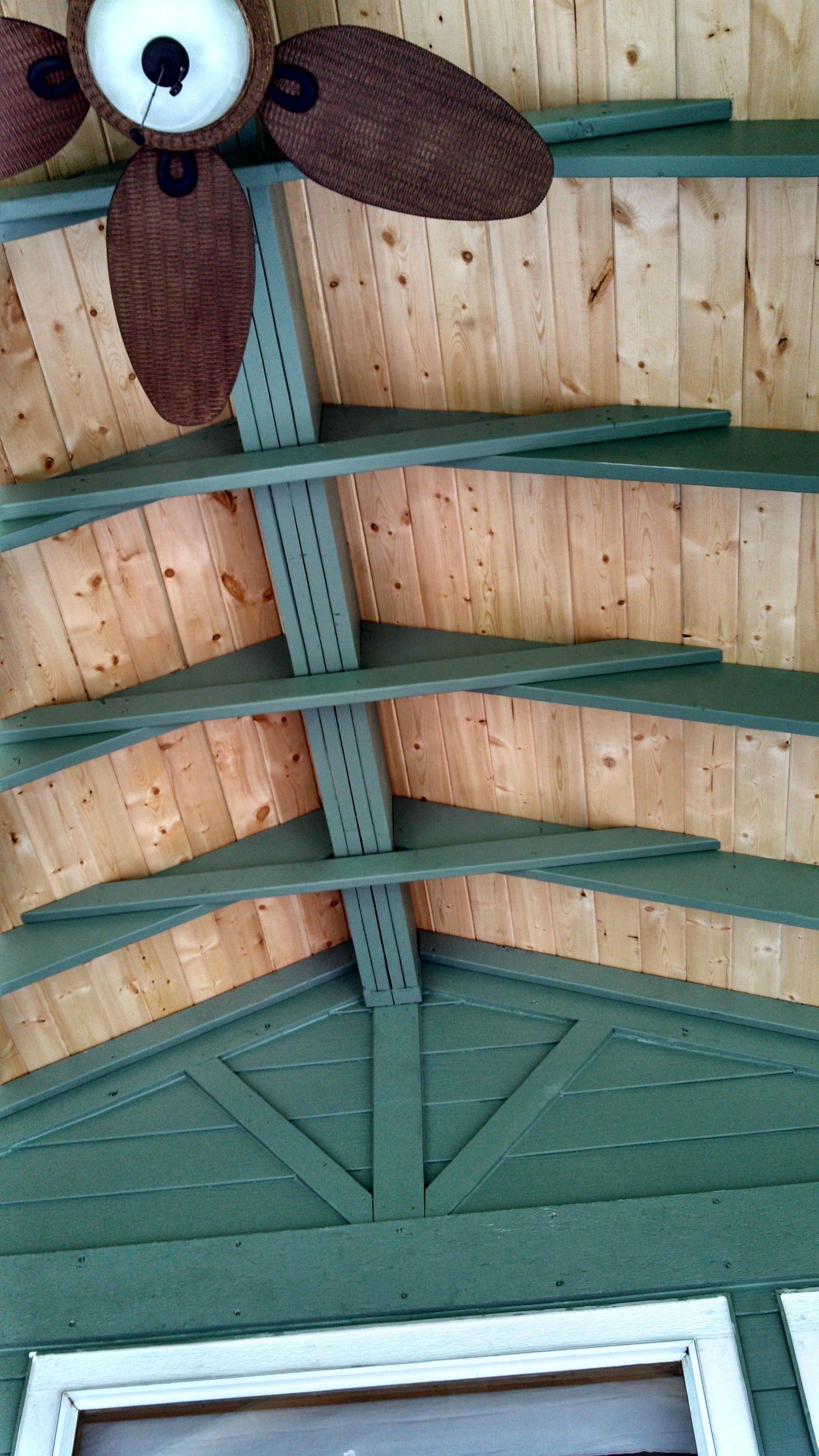 Wooden porch ceiling with green beams and a brown ceiling fan against wood paneling.