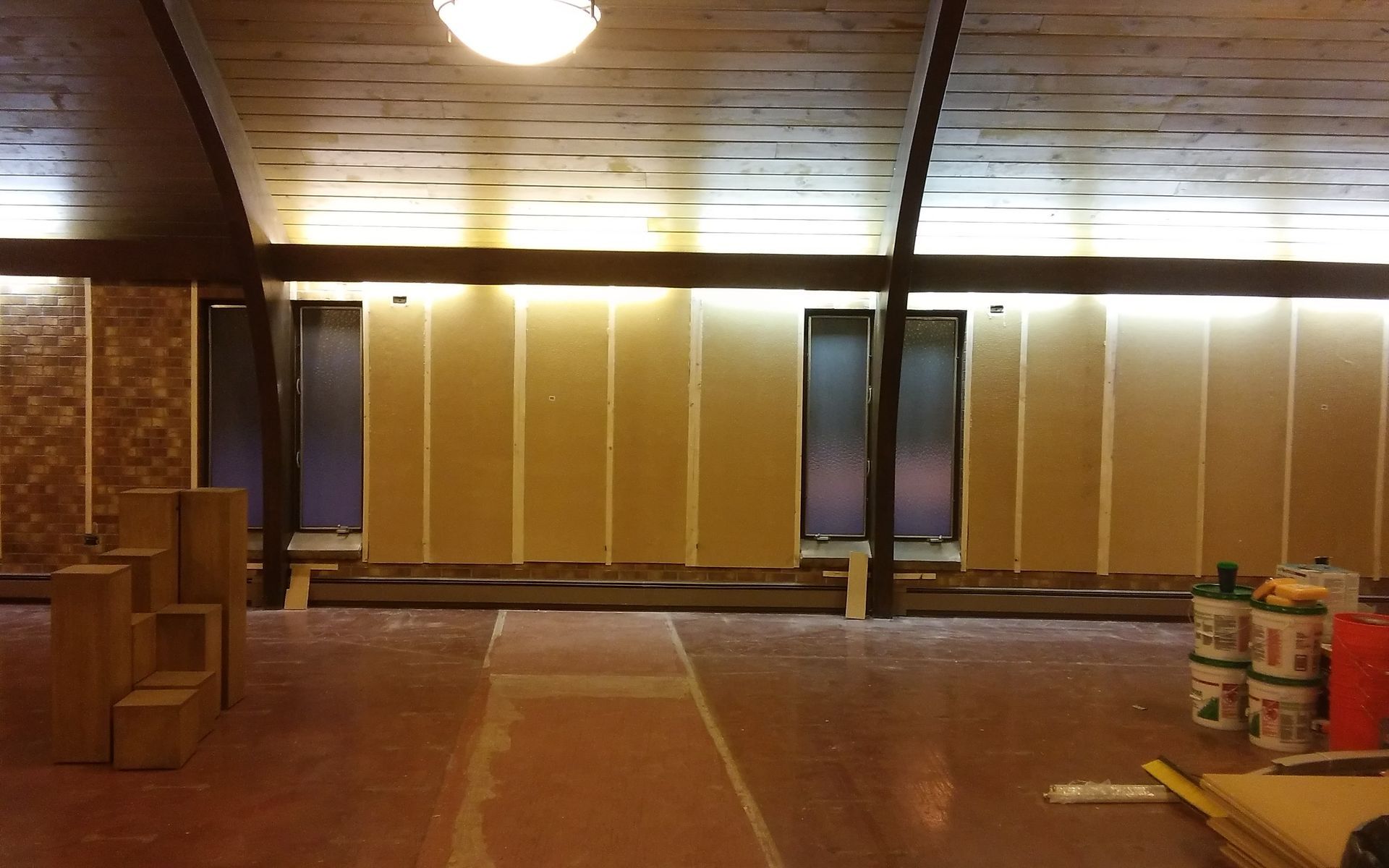 Empty unfinished room with wood-paneled walls, ceiling lights, and stacked paint buckets on the right.