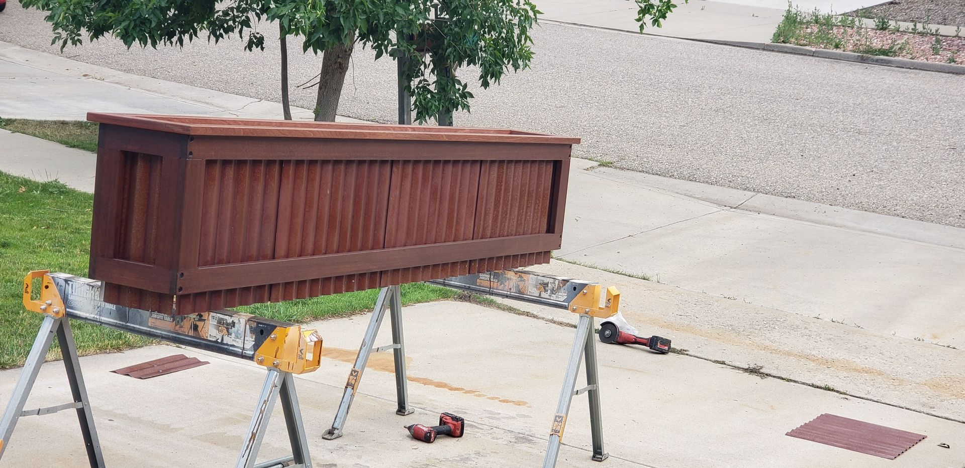 Brown wooden planter box on sawhorses beside a street, with a small tree behind it.