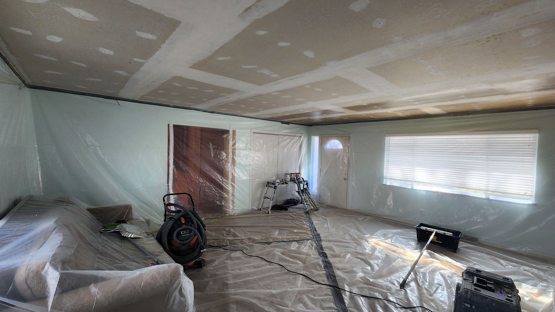 Construction room with taped-off ceiling, covered floor, tools, and a window letting in light.