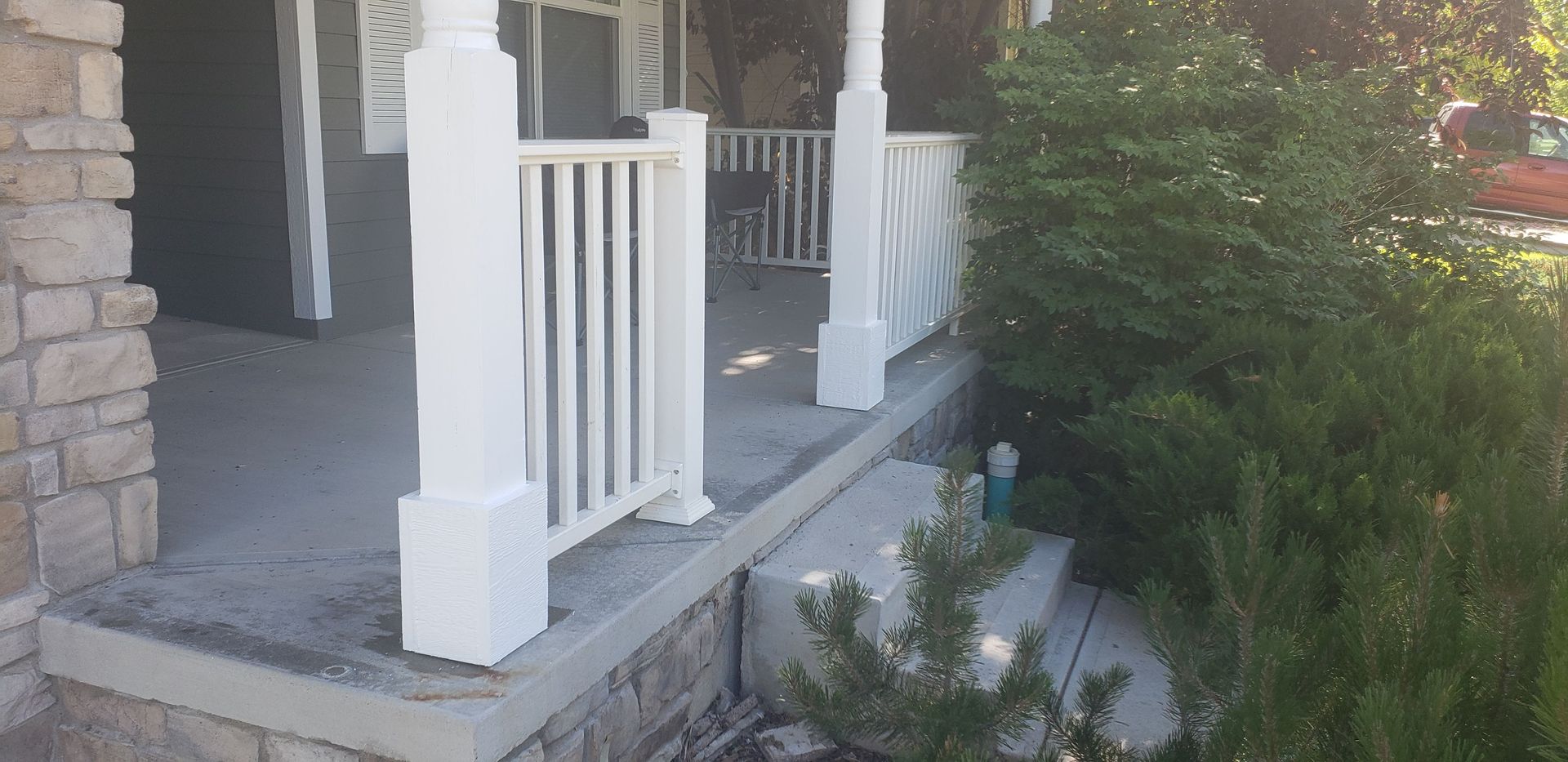Front porch with white railings and steps beside stone siding and green shrubs.