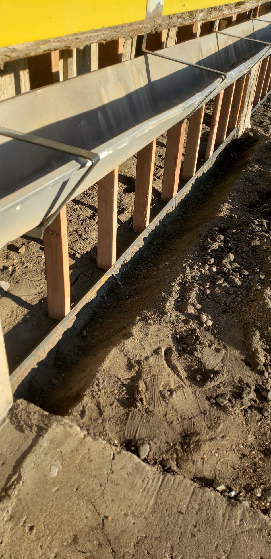 Wooden formwork and a metal chute running along a trench beside a yellow wall under construction.