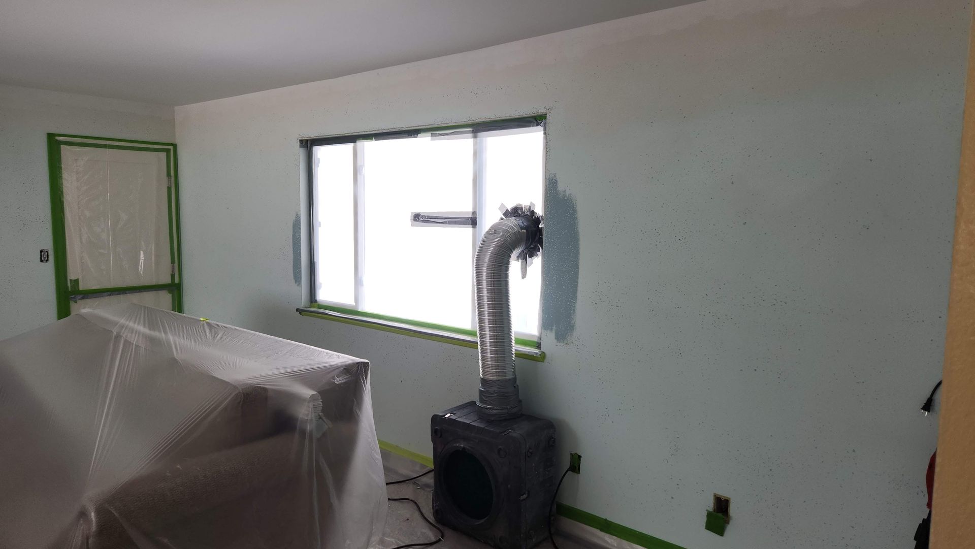 Partially painted room with a window, green trim, and a stove with vent pipe; furniture covered in plastic.