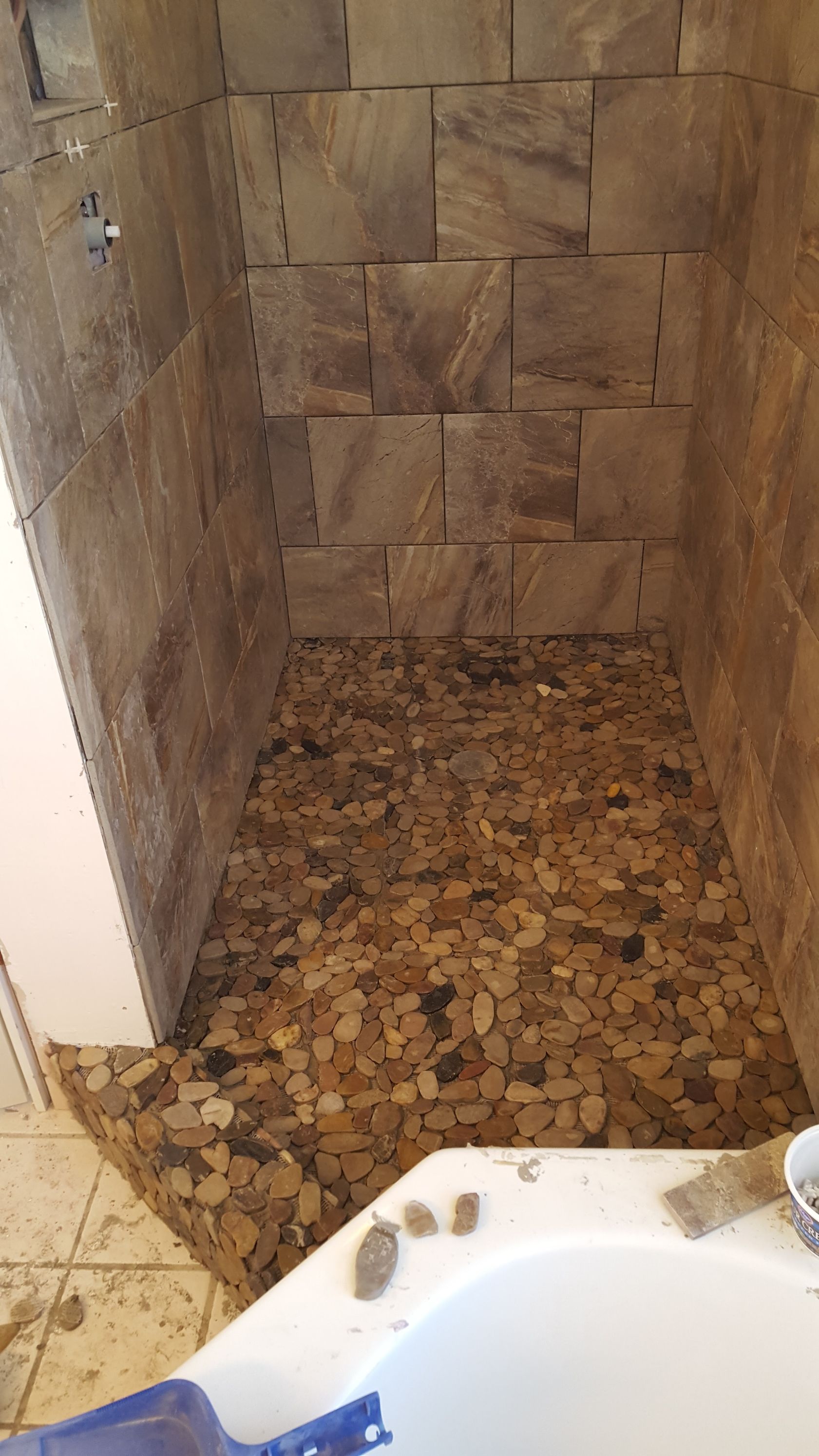 Narrow tiled shower alcove with stone wall and pebble floor beside a white tub edge.