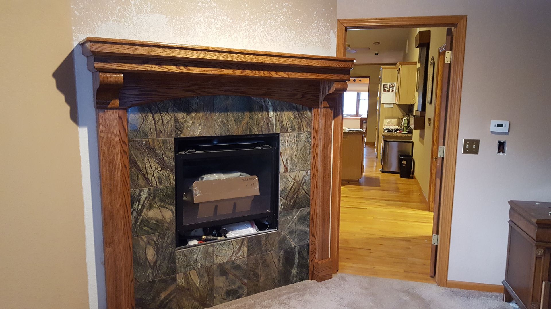 Living room with stone fireplace and wood mantel, looking into a brightly lit hallway and kitchen.