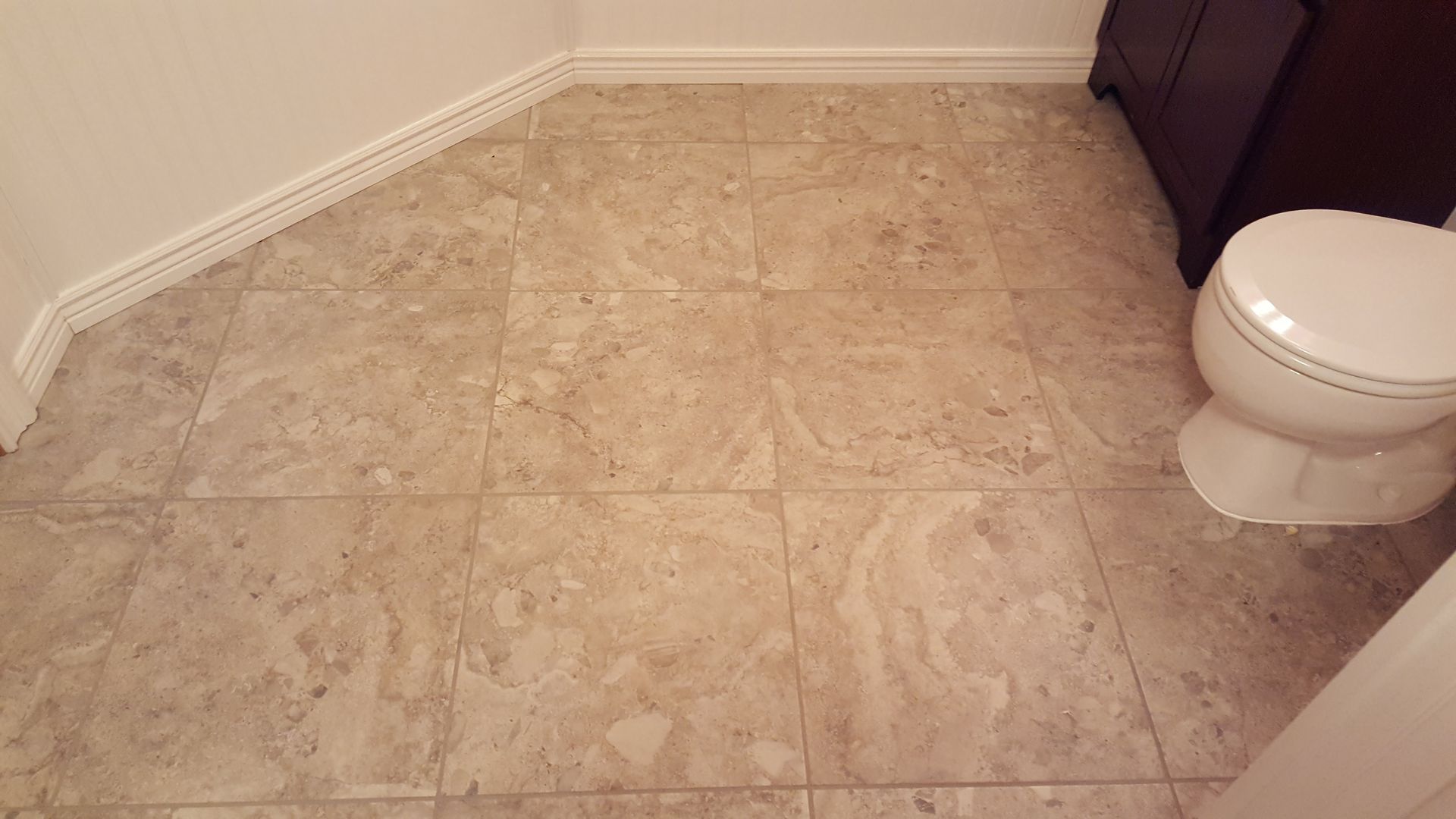 Tiled bathroom floor with white toilet and beige walls, shown from a corner view.