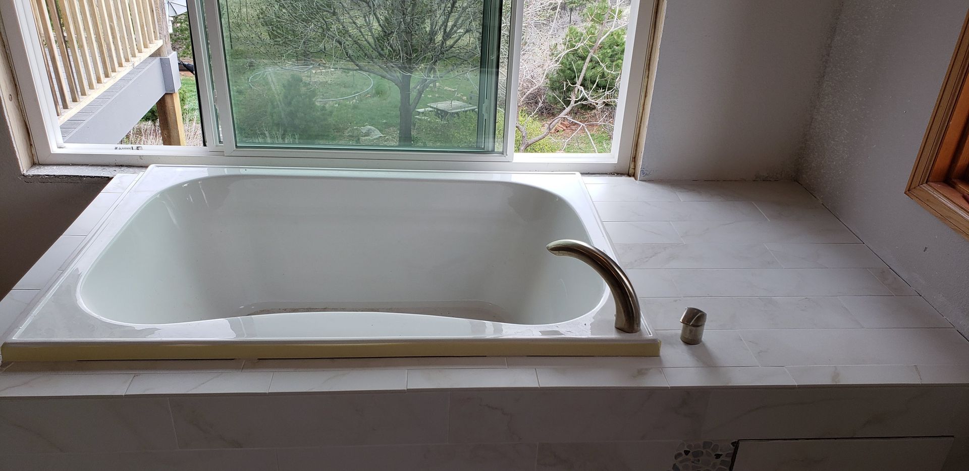 White bathtub by a window overlooking greenery, with a faucet on the right and a tiled surround.