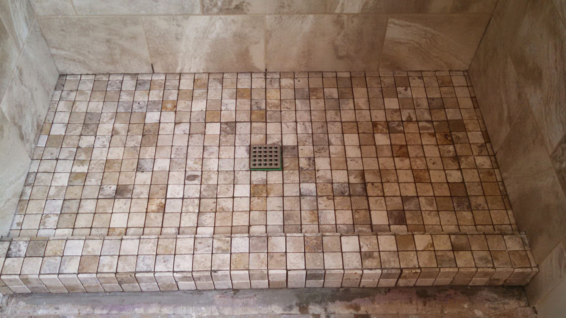 Small tiled shower floor with a central drain and beige stone walls.