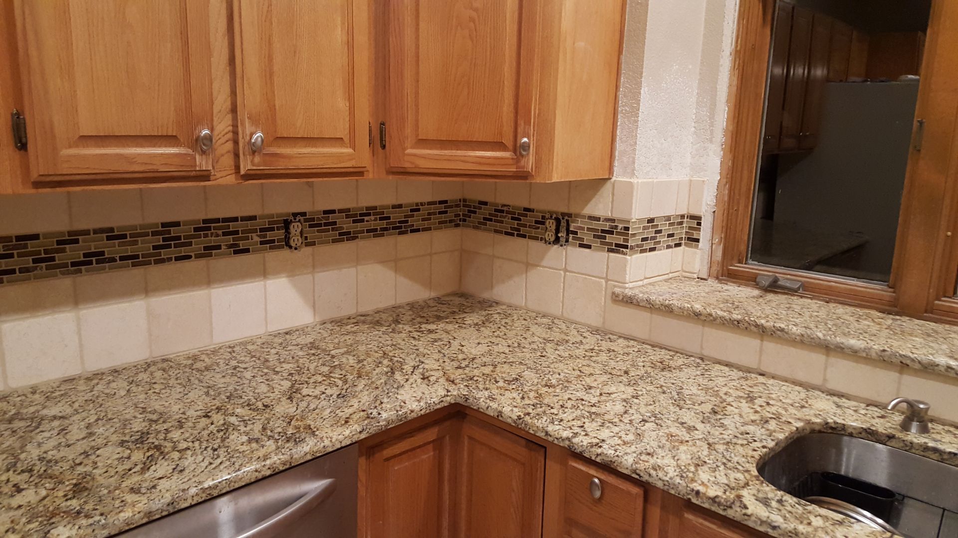 Corner kitchen countertop with beige granite, wood cabinets, tiled backsplash, and window above the sink.