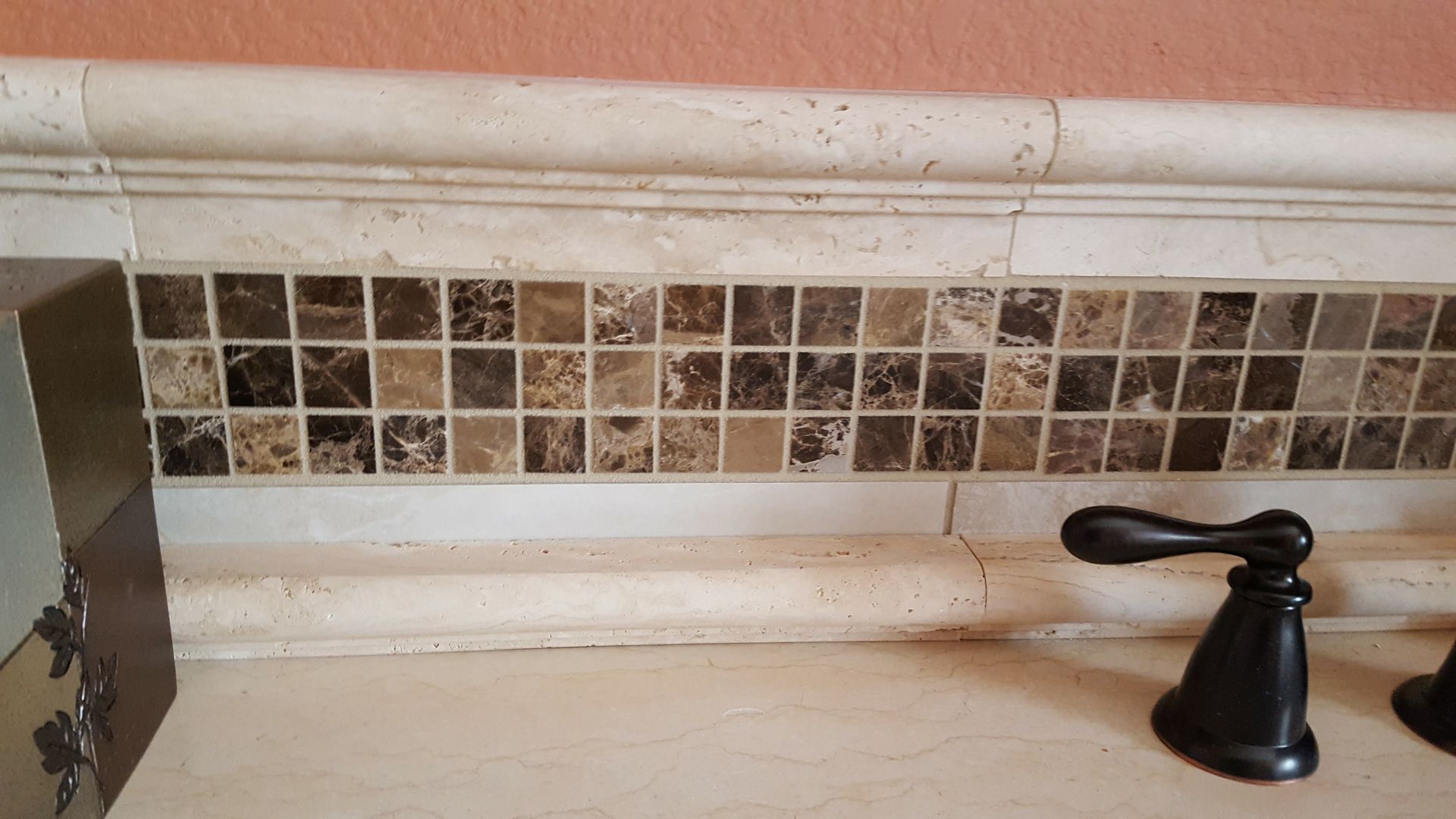 Bathroom sink backsplash with beige tile mosaic and black faucet on the right.