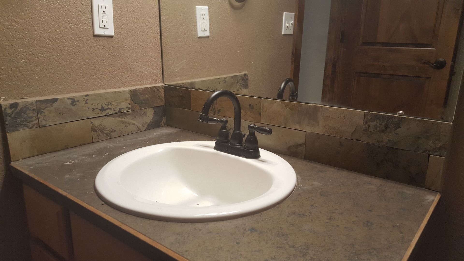 Bathroom vanity with a white sink, dark faucet, and granite countertop against a tan wall.